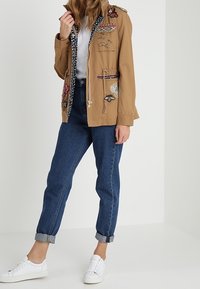 Brown lightweight jacket with embroidered floral designs and a zip closure. Paired with rolled-up dark blue jeans and white sneakers.