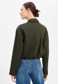 Donkergroene blouse met een ontspannen pasvorm, lange mouwen en een kraag, gecombineerd met lichtblauwe jeans met hoge taille. Gladde stoftextuur.