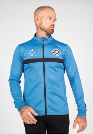 Blauwe sportjack met zwarte horizontale streep en logo, met een hoge kraag, ritssluiting en lange mouwen. Glad, rekbaar materiaal.