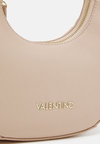 Bolsa bege com uma superfície texturada suave, detalhes em zíper dourado e logotipo "VALENTINO" em relevo na parte da frente. Forma circular.
