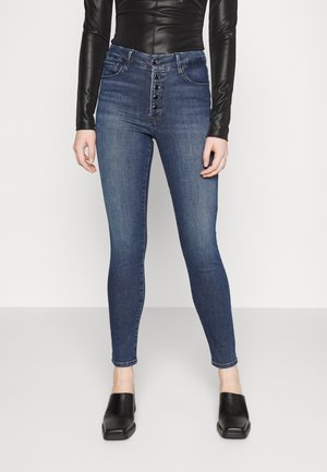 Woman wearing high-waisted blue skinny jeans with button closure and black square-toe slip-on shoes standing against a plain background.