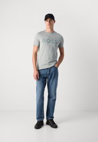 Camiseta de algodón verde claro con "BOSS" impreso en letras grandes. Combinada con jeans de denim azul y zapatillas negras. Los accesorios incluyen una gorra negra.