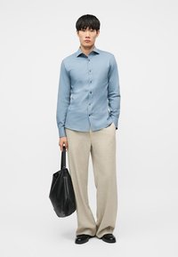 Άνδρας που φοράει ανοιχτόμπλε πουκάμισο με κουμπιά και μπεζ παντελόνι φαρδύ, κρατώντας με το ένα χέρι μια μαύρη δερμάτινη τσάντα ώμου, στέκεται σε λευκό φόντο.