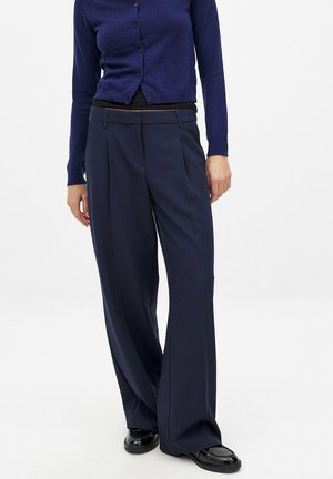 Navy wide-leg trousers with pleats, made of smooth fabric. Paired with a dark blue long-sleeve cardigan and glossy black shoes.