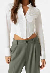 Donna con una camicia bianca cropped e bottoni, maniche lunghe, e pantaloni olive verdi a vita alta, in piedi contro uno sfondo neutro.