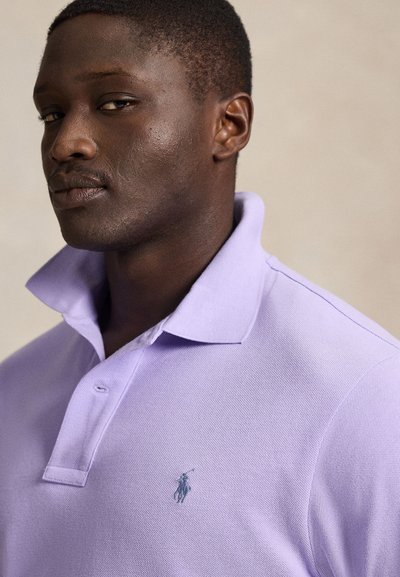 Κοντινό πλάνο ενός άντρα που φοράει ένα πασχαλιά polo πουκάμισο με ανασηκωμένο κολάρο, σε έναν απλό ουδέτερο φόντο.