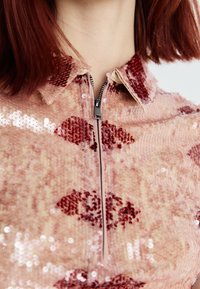 T-shirt zippé avec une base rose clair orné de motifs en sequins rouges. Texture douce et col visible, doté d'une fermeture éclair métallique.