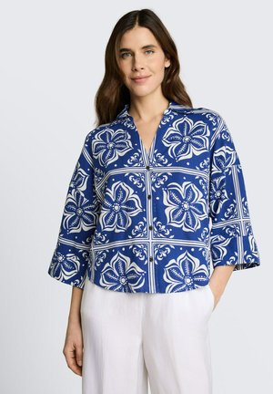 Femme portant un chemisier bleu boutonné à motifs floraux blancs et manches trois-quarts, associé à un pantalon blanc, posant avec une main dans la poche.