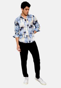 Camicia button-up in blu, beige e un motivo geometrico nero, abbinata a pantaloni neri e sneakers bianche. La camicia presenta maniche arrotolate.