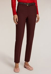 Bordeauxkleurige op maat gemaakte broek met een slim fit, voorzien van een zwarte riem met een gouden gesp. Gecombineerd met roze versierde flats.
