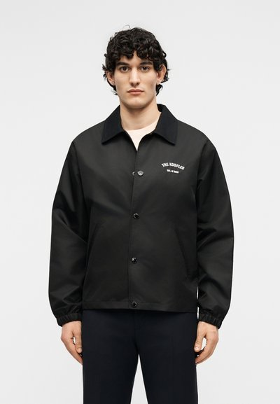 Νεαρός άνδρας που φοράει μαύρο σακάκι με κουμπιά και το λογότυπο «The Kooples» στο στήθος, στέκεται μπροστά σε ένα απλό φωτεινό φόντο.