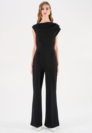 Combinaison noire avec un haut drapé à une épaule et un pantalon à jambes larges. Présente une taille cintrée et une texture de tissu lisse.