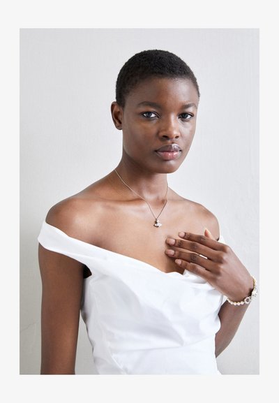 Silver necklace with a small pendant, worn on bare skin accentuated by an off-the-shoulder white dress. Smooth texture and elegant design.