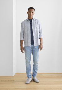 Cămașă cu dungi albă și bleumarin, purtată peste un tricou bleumarin. Blugi deschiși și adidași gri completează ținuta. Fundal cu parchet din lemn.