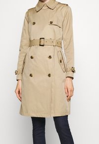 Beige trenčkot s dvojitými knoflíky, páskem v pase, klopovými kapsami a nastavitelými pásky na rukávech, nošený přes tmavé kalhoty.