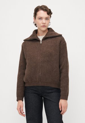 Ruda zip-up megztinis su aukštu apykaklė, mezginio tekstūra, briaunoti rankogaliai ir apatinė dalis. Dėvimas virš šviesios spalvos marškinėlių, derinamas su tamsiomis džinsomis.