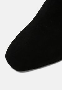 Bottine en daim noir à bout pointu, texture lisse et silhouette épurée, affichant des détails de couture le long des bords.