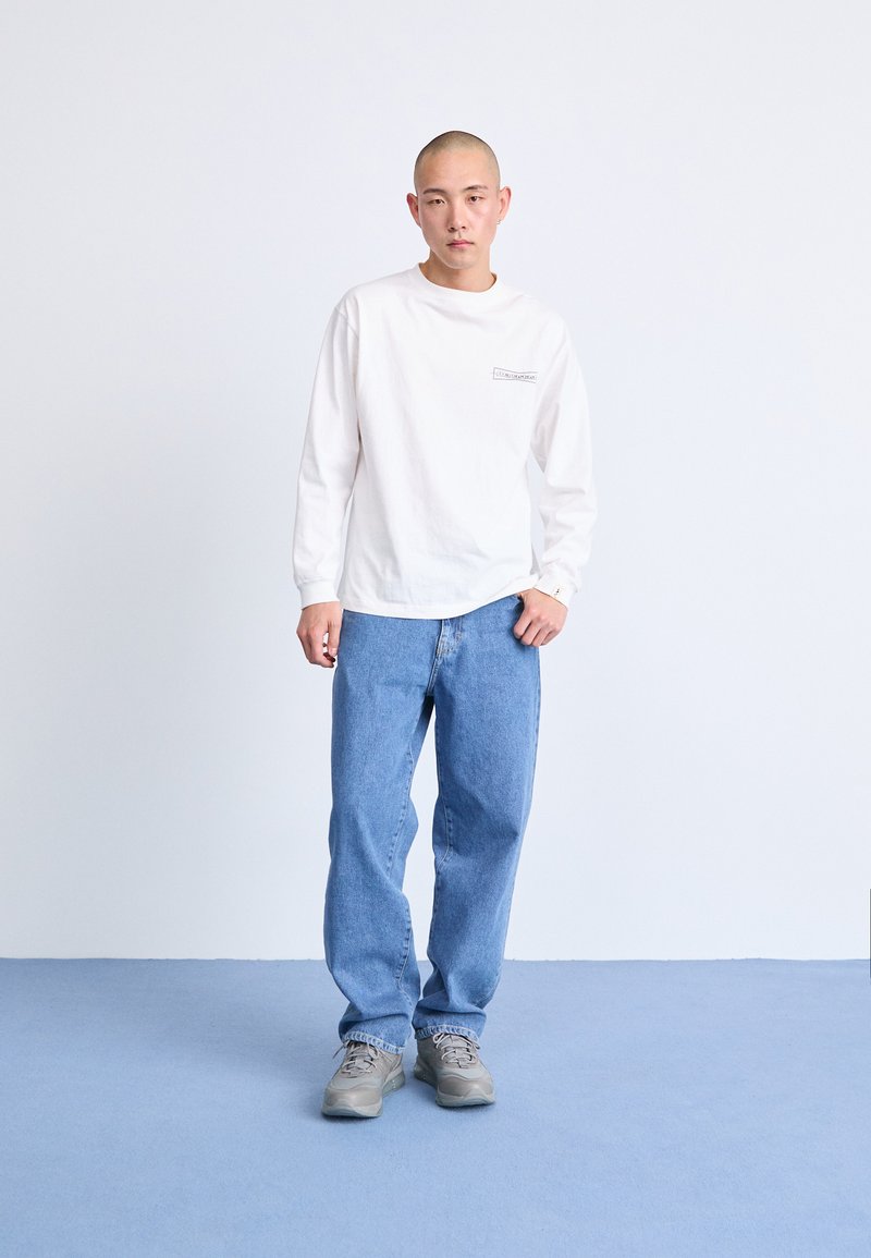 Weißes Langarmhemd mit einem Brustlogo, kombiniert mit hellblauen, locker sitzenden Jeans und grauen Turnschuhen. Einfaches Design, neutrale Farben.