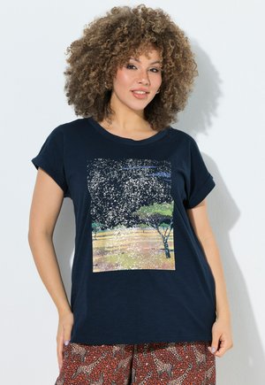 Kvinde med krøllet hår iført en marineblå T-shirt med print af et stjerneklar natlandskab og mønstrede bukser med girafmotiver.