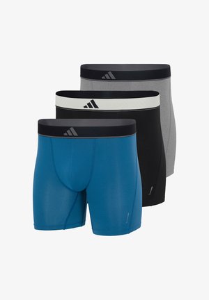 Oberkörperfreier Mann trägt blaue figurbetonte Boxershorts mit schwarzem Bund, steht barfuß vor einem schlichten weißen Hintergrund.