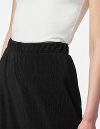 Jupe noire avec un design texturé et plissé, dotée d'une large ceinture élastique, associée à un haut blanc ajusté. Un angle en gros plan met en valeur les détails.
