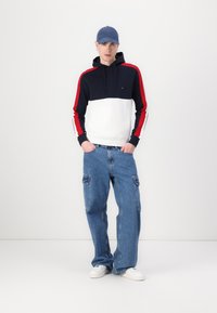 Hooded sweatshirt with navy, white, and red color blocks, paired with loose blue cargo jeans and white sneakers. The cap is navy.