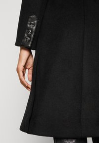 Zwarte wollen jas met een mouw met drie zwarte knopen op een leren manchet, gedragen door een persoon met een zichtbaar hand aan de zijkant.