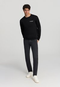 Felpa nera con collo rotondo e scritta bianca sul petto, abbinata a jeans scuri e sneakers bianche. Tessuto morbido, vestibilità casual.