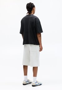 Sort, oversized kortærmet skjorte med synlige stikninger, parret med lysegrå shorts der går til knæene. Sportsskoene har blå detaljer.