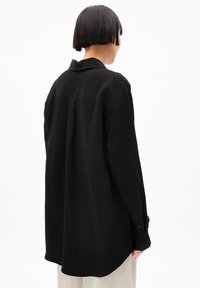 Schwarzes, übergroßes Hemd mit Kragen, langen Ärmeln und Knopfmanschetten. Verfügt über eine Rückenpasse mit dezenten Falten und besteht aus weichem Stoff.