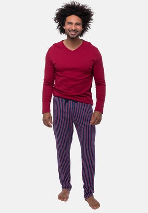 Man met krullend haar die lacht, draagt een rood shirt met lange mouwen en blauw gestreepte pyjamabroek, staat blootsvoets op een effen achtergrond.