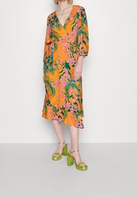 Kwiatowa sukienka midi w intensywnym pomarańczowym kolorze, z różowymi kwiatami i zielonymi liśćmi. Dekolt w kształcie litery V, rękawy o długości trzy czwarte oraz falbaniasty dół.