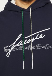 Tmavě modrá mikina s kapucí s bílým logem Lacoste a motivem krokodýla, se zelenobílými šňůrkami s texturou. Měkká látka, standardní střih.