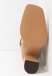 Bottine en daim beige avec un bout légèrement pointu et un talon empilé, dotée d'une semelle en caoutchouc lisse à rainures horizontales.