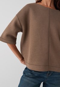 Sweatshirt marron oversize avec un col large et des manches courtes. Fait en un tissu doux, avec des coutures visibles et une coupe décontractée.