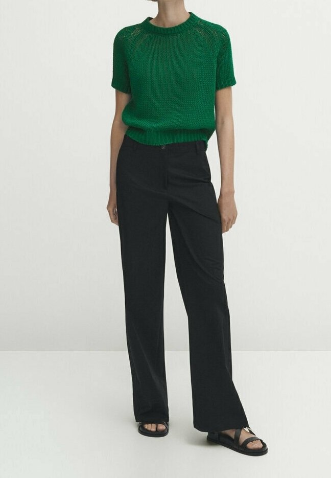 Personne portant un pull vert en tricot à manches courtes, un pantalon noir large taille haute, et des sandales noires à bouts ouverts, debout sur un sol blanc.