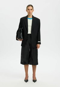 Veste noire sur mesure, haut à blocs de couleur blanc et bleu, culottes en cuir noir, tenant un sac à main noir avec une poignée circulaire, et escarpins noirs.