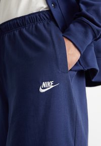 Tmavě modré tepláky Nike s elastickým pasem a bílým logem na nohavici. Tkanina vypadá hladce a jemně.