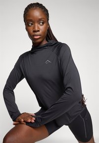 Mustast spordist pika varrukaga kapuutsiga särk, kus rinnal on peen logo ja sile tekstuur, kombineeritud mustade lühikeste pükstega.