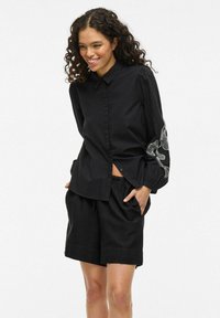 Camicia nera con colletto, maniche a sbuffo e ricami decorativi bianchi sulla manica sinistra; abbinata a pantaloni corti neri.