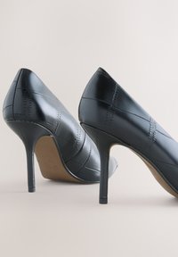 Paire de chaussures à talons hauts en cuir noir avec motif carré cousu, talons fins et semelles marron clair sur fond neutre.