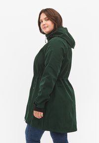 Bosgroene jas met capuchon, getailleerde taille en ritszakken. Gemaakt van zacht materiaal met een gladde textuur en elastische manchetten.
