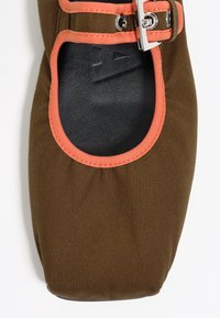 Zapato de lona marrón con un ribete naranja, puntera redonda y un detalle de hebilla plateada para sujetar en el lateral. El interior tiene un forro negro.
