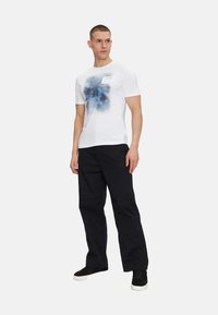 Camiseta blanca con patrón abstracto azul, de manga corta y cuello redondo; combinada con pantalones negros de corte holgado y zapatillas negras.
