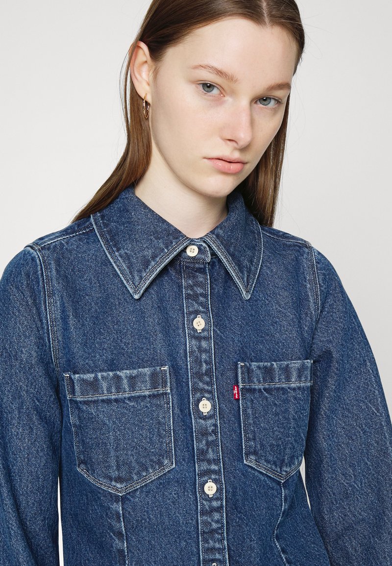 Camisa de denim en azul oscuro, con collar puntiagudo, botones frontales y dos bolsillos en el pecho. Tejido texturizado con acentos desgastados.