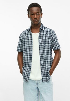 Jeune homme aux cheveux courts et bouclés portant une chemise à manches courtes à carreaux bleu et blanc par-dessus un t-shirt blanc et un jean bleu clair, les mains dans les poches.