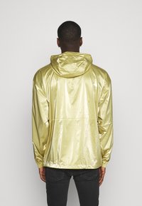 Hombre con una chaqueta metálica dorada brillante con capucha y jeans negros, visto desde atrás contra un fondo liso claro.