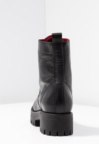 Černá kožená bota s robustní podrážkou, viditelným zadním švem, poutkem na vtažení a červeným vnitřním podšívkou, zobrazená ze zadní strany na bílé ploše.