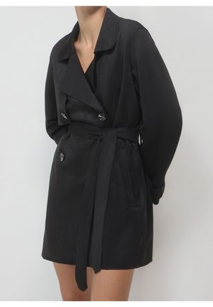 Kvinde iført en sort dobbeltradet trenchcoat med brede revers og et bælte bundet i taljen mod en ensfarvet baggrund.