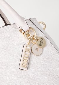 Borsa bianca con texture, dotata di hardware dorato, due portachiavi con loghi del marchio e un'etichetta decorativa con testo in rilievo.